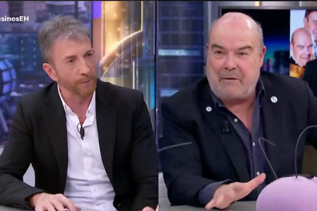 Pablo Motos y Antonio Resines conversan en 'El Hormiguero', ambos visten trajes oscuros y están sentados en una mesa con un fondo de estudio iluminado.