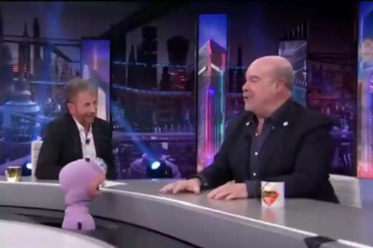 Pablo Motos y Antonio Resines conversan en 'El Hormiguero' con luces de colores y un muñeco morado en la mesa. Pablo Motos y Antonio Resines conversan en 'El Hormiguero' con luces de colores y un muñeco morado en la mesa.