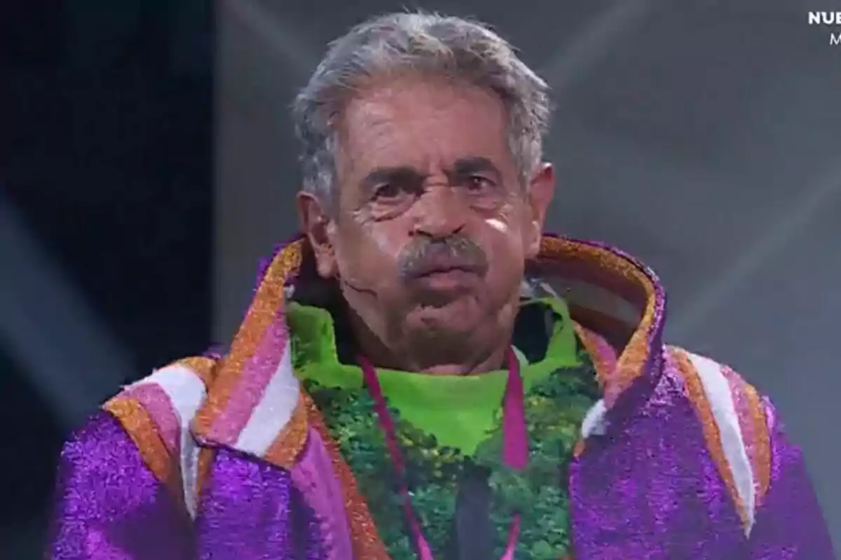 Miguel Ángel Revilla con bigote lleva un colorido disfraz con tonos morados y naranjas en un escenario iluminado en Mask Singer. Miguel Ángel Revilla con bigote lleva un colorido disfraz con tonos morados y naranjas en un escenario iluminado en Mask Singer.