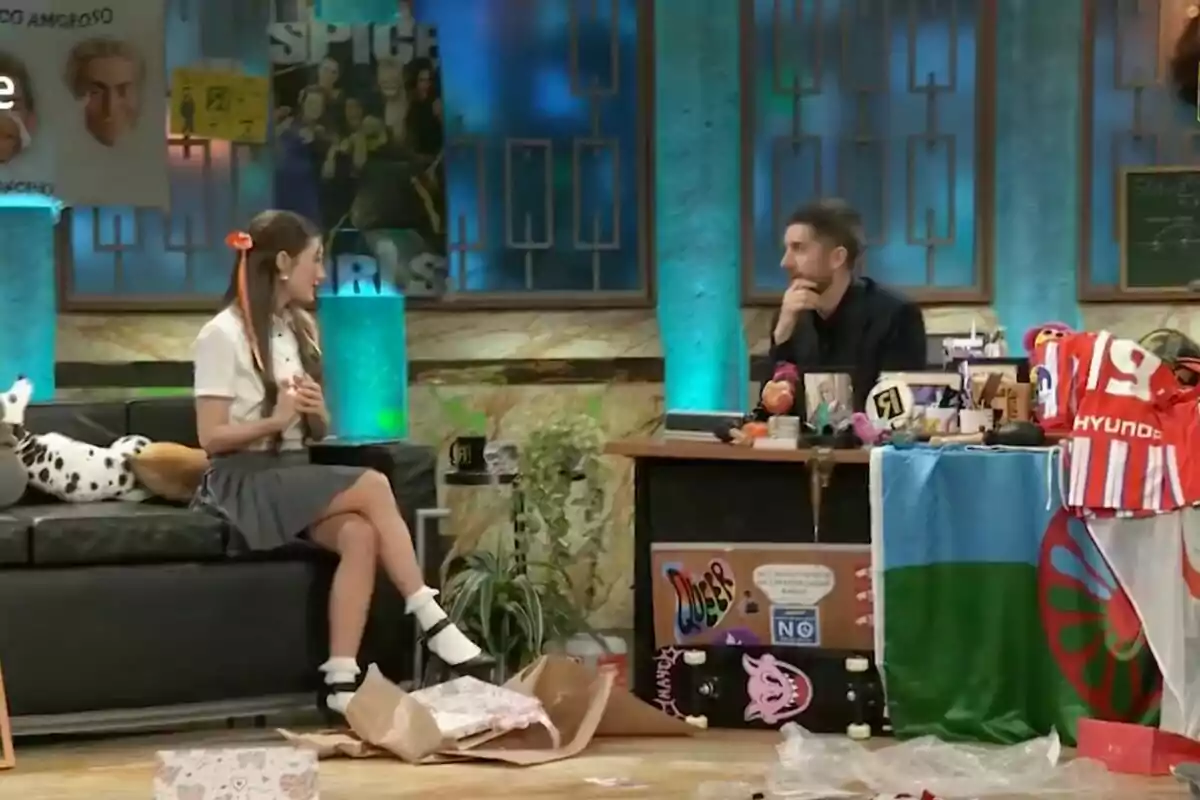 Yami Safdie con uniforme escolar está sentada en un sofá frente a David Broncano en un escritorio lleno de objetos variados, en un set de televisión decorado con carteles y luces azules en 'La Revuelta'. Yami Safdie con uniforme escolar está sentada en un sofá frente a David Broncano en un escritorio lleno de objetos variados, en un set de televisión decorado con carteles y luces azules en 'La Revuelta'.