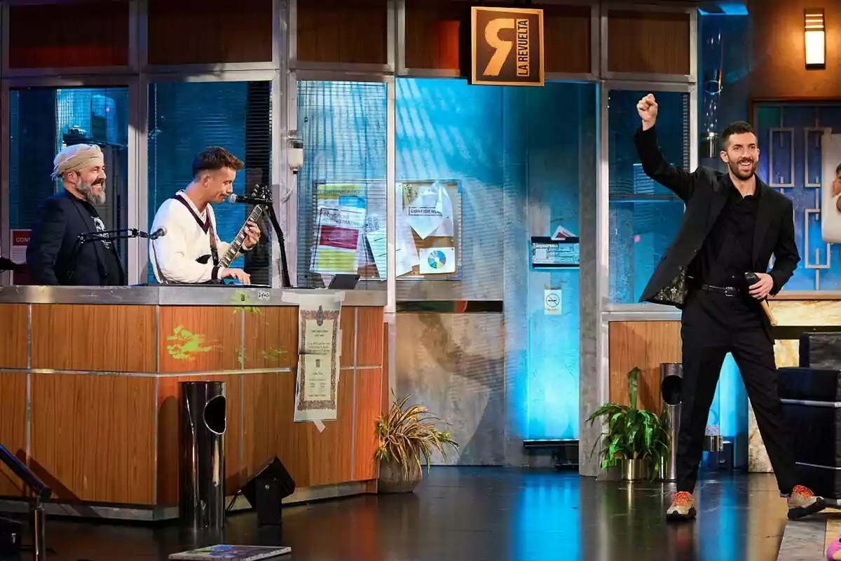 David Broncano con traje negro levanta el puño mientras Ricardo Castella y Grison están detrás de un mostrador, uno de ellos tocando una guitarra, en un set de televisión con un letrero que dice David Broncano con traje negro levanta el puño mientras Ricardo Castella y Grison están detrás de un mostrador, uno de ellos tocando una guitarra, en un set de televisión con un letrero que dice