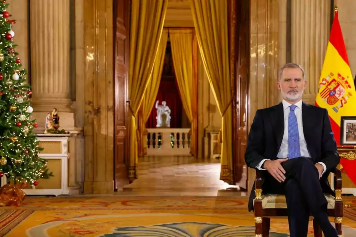 Felipe VI sentado en una silla en un elegante salón decorado con un árbol de Navidad y una bandera de España. Felipe VI sentado en una silla en un elegante salón decorado con un árbol de Navidad y una bandera de España.