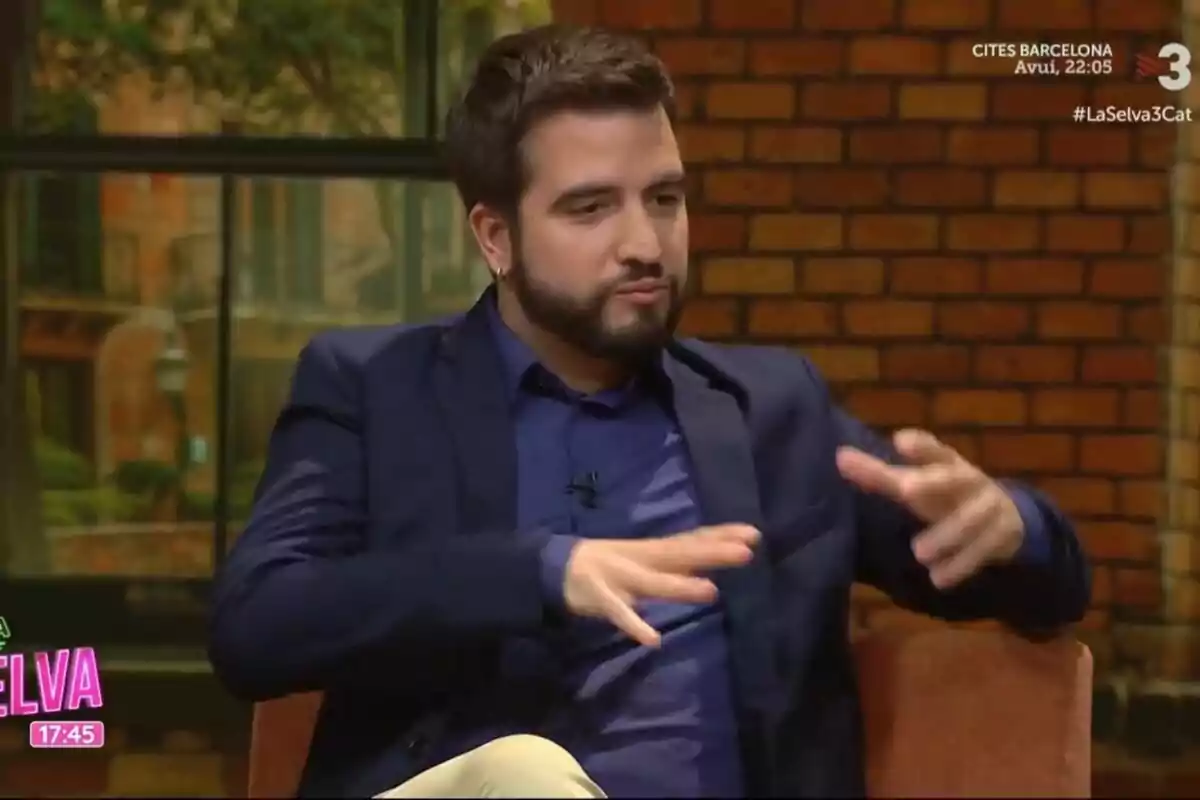 Un home amb barba i jaqueta blava gesticula mentre està assegut en un set de televisió amb fons de maons.