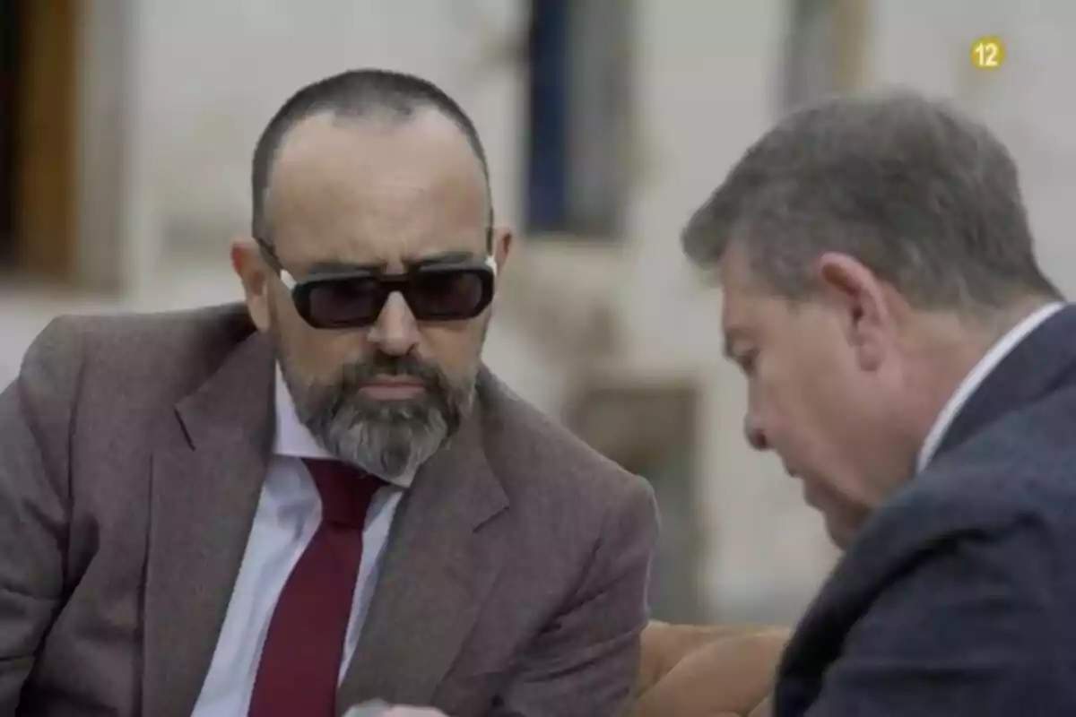 Risto Mejide y Emiliano García-Page conversan mientras uno de ellos lleva gafas de sol y traje marrón.
