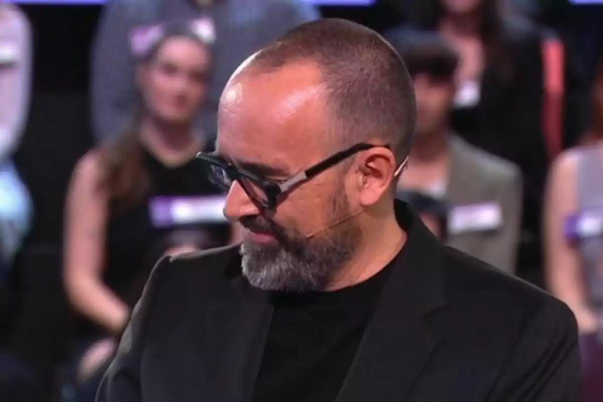 Risto Mejide con gafas y barba en Demos: El Gran Sondeo, mirando hacia abajo, con personas desenfocadas en el fondo. Risto Mejide con gafas y barba en Demos: El Gran Sondeo, mirando hacia abajo, con personas desenfocadas en el fondo.