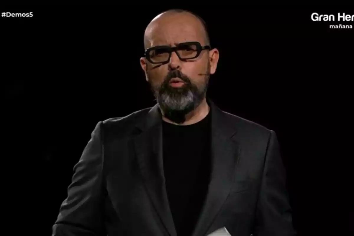 Risto Mejide con barba y gafas en un escenario oscuro, vistiendo un traje negro en Demos: El Gran Sondeo. Risto Mejide con barba y gafas en un escenario oscuro, vistiendo un traje negro en Demos: El Gran Sondeo.