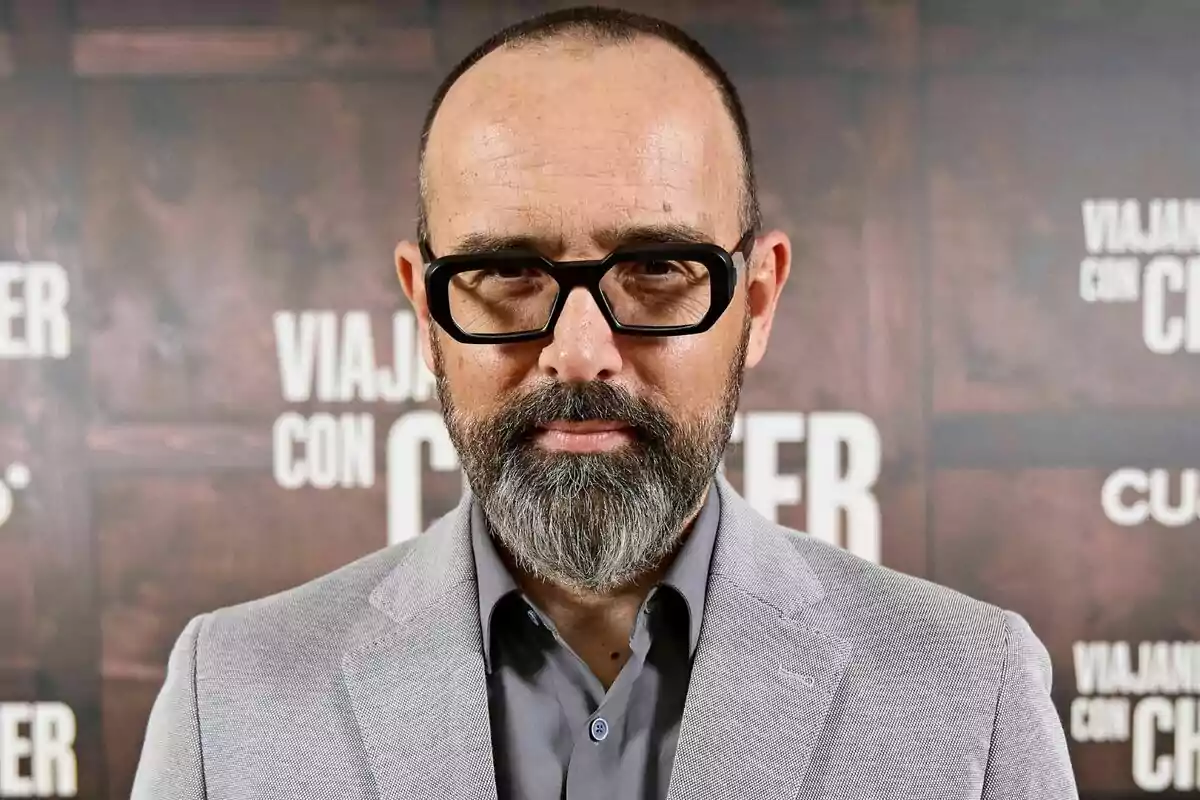 Risto Mejide amb barba i ulleres negres, vestit amb un vestit gris, davant d'un fons amb text borros de Viajando con Chester.
