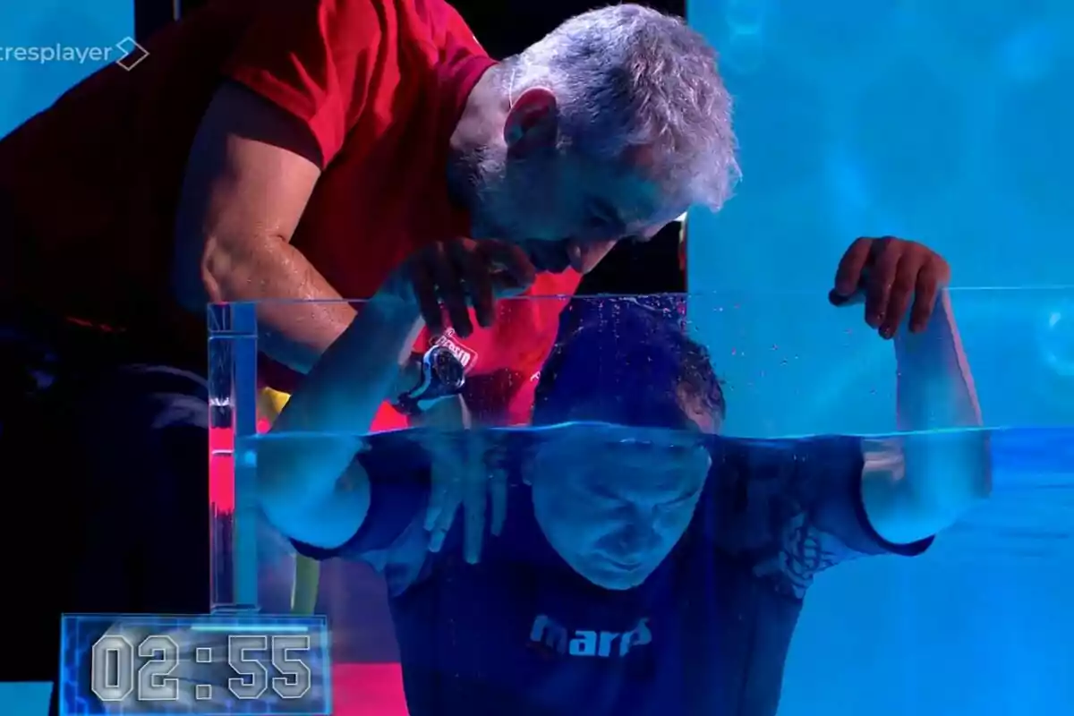 Un hombre con camiseta roja observa a Roberto Brasero que está sumergido en un tanque de agua en El Desafío, mientras un cronómetro en la esquina inferior izquierda muestra 02:55. Un hombre con camiseta roja observa a Roberto Brasero que está sumergido en un tanque de agua en El Desafío, mientras un cronómetro en la esquina inferior izquierda muestra 02:55.