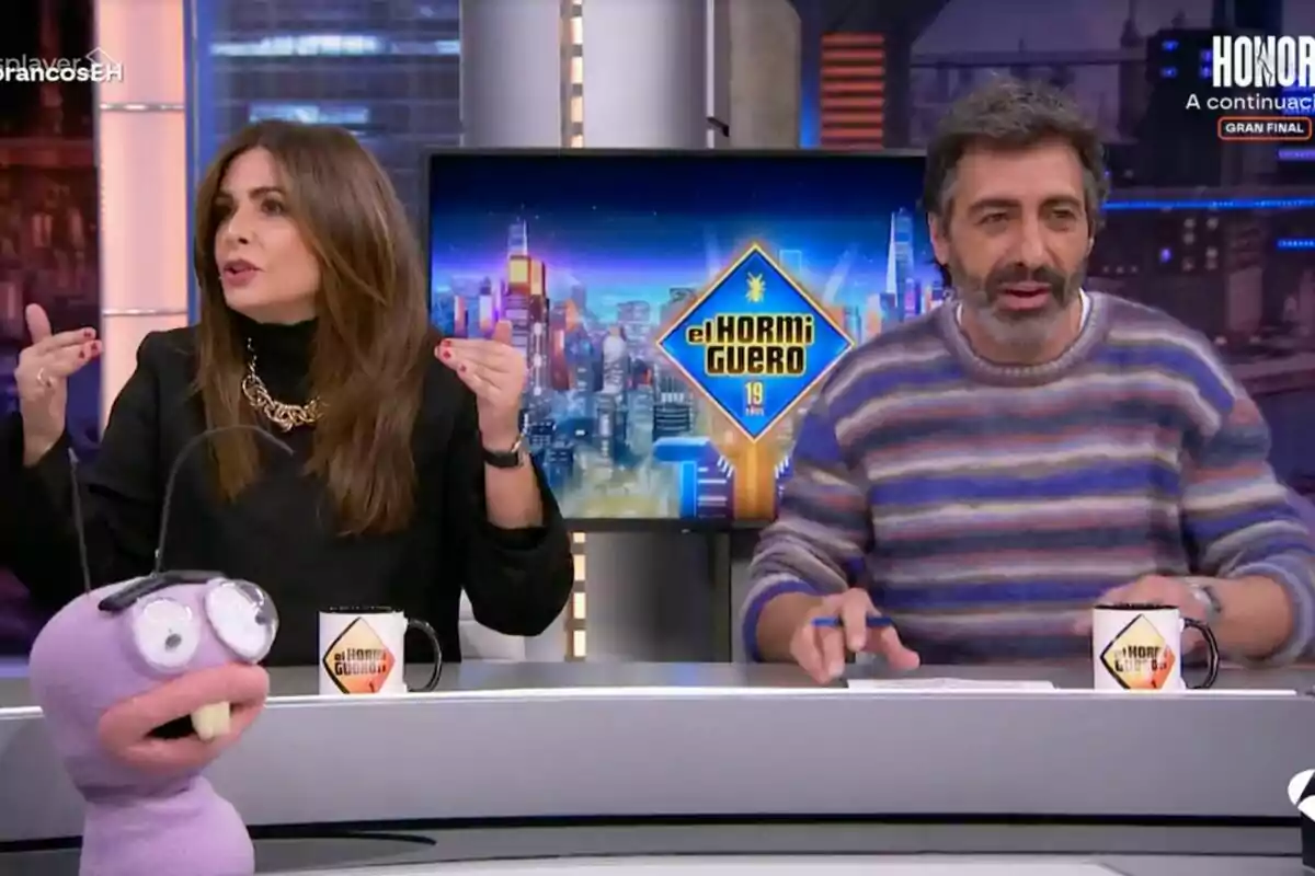 Nuria Roca y Juan del Val sentadas en un set de televisión con una marioneta y tazas con el logo de "El Hormiguero" en la mesa.
