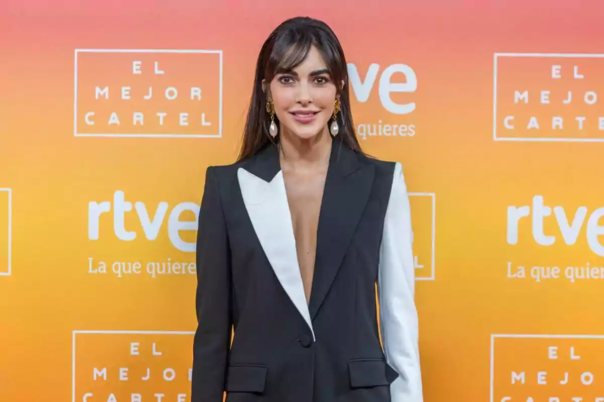 Rocío Muñoz con un traje de chaqueta negro y blanco posa frente a un fondo naranja con logotipos de Rocío Muñoz con un traje de chaqueta negro y blanco posa frente a un fondo naranja con logotipos de