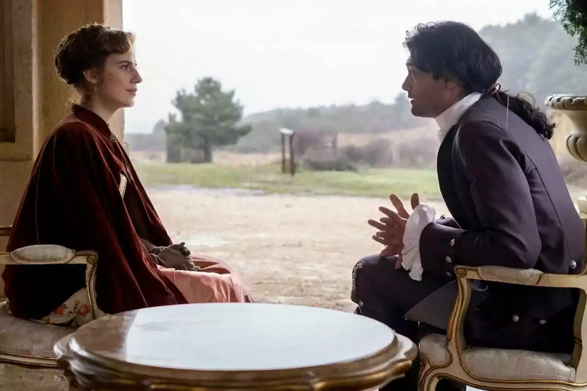 Rocío Súarez de Puga y Marco Pernas vestidos con ropa de época están sentados frente a frente en sillas elegantes, en un entorno al aire libre con un paisaje natural de fondo en Valle Salvaje. Rocío Súarez de Puga y Marco Pernas vestidos con ropa de época están sentados frente a frente en sillas elegantes, en un entorno al aire libre con un paisaje natural de fondo en Valle Salvaje.