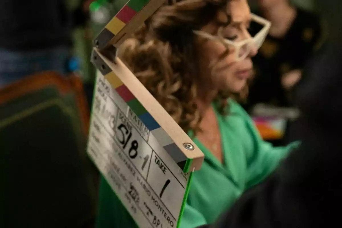 Secuencia del rodaje de 'Nails'. Una persona con gafas grandes y cabello rizado está en un set de filmación, mientras una claqueta de cine se encuentra en primer plano. Secuencia del rodaje de 'Nails'. Una persona con gafas grandes y cabello rizado está en un set de filmación, mientras una claqueta de cine se encuentra en primer plano.