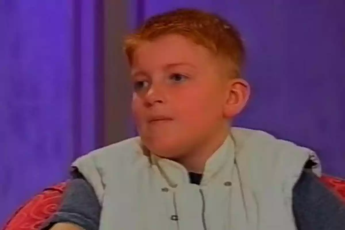 Rory Callum Sykes pèl-roig amb expressió pensativa està assegut en un sofà amb fons morat.