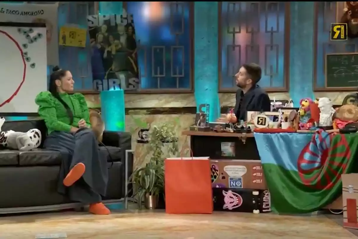 Rosa López con un atuendo verde está sentada en un sofá negro mientras conversa con David Broncano que está detrás de un escritorio lleno de objetos variados en un set de televisión decorado con carteles y luces azules en 'La Revuelta'. Rosa López con un atuendo verde está sentada en un sofá negro mientras conversa con David Broncano que está detrás de un escritorio lleno de objetos variados en un set de televisión decorado con carteles y luces azules en 'La Revuelta'.