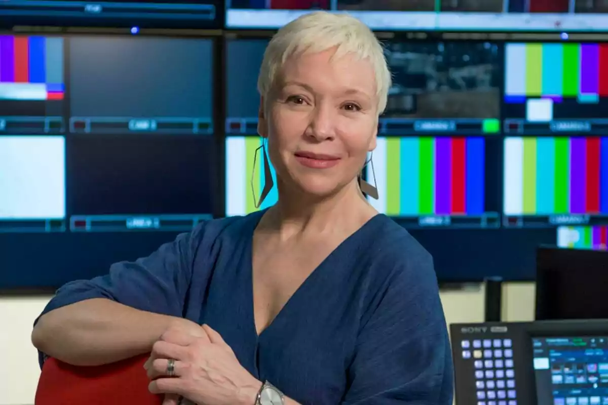 Fotografía de Rosa María Molló de cabello corto y rubio con pendientes largos, vestida con blusa azul, posando frente a múltiples pantallas de colores en un estudio de televisión en RTVE Fotografía de Rosa María Molló de cabello corto y rubio con pendientes largos, vestida con blusa azul, posando frente a múltiples pantallas de colores en un estudio de televisión en RTVE