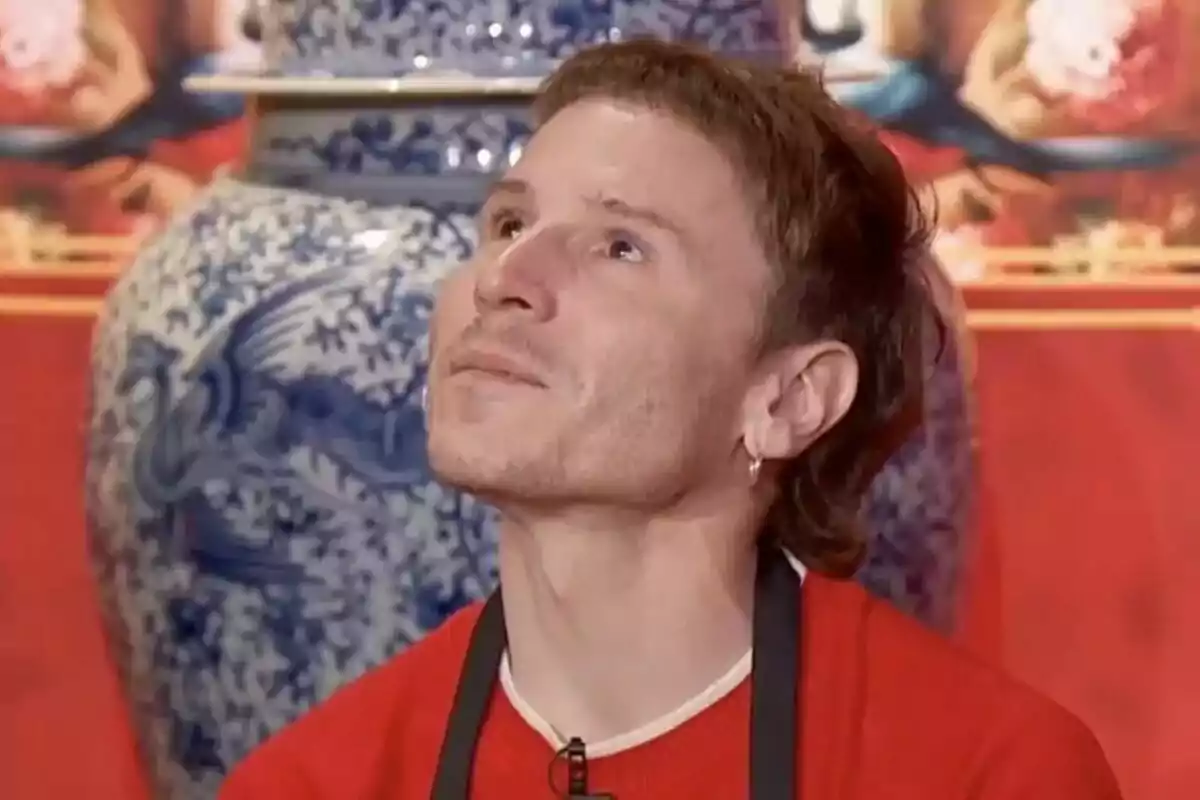 Rubén Ochandiano con una camiseta roja y un delantal negro mira hacia arriba, con un fondo que incluye un jarrón azul y blanco en MasterChef Celebrity. Rubén Ochandiano con una camiseta roja y un delantal negro mira hacia arriba, con un fondo que incluye un jarrón azul y blanco en MasterChef Celebrity.