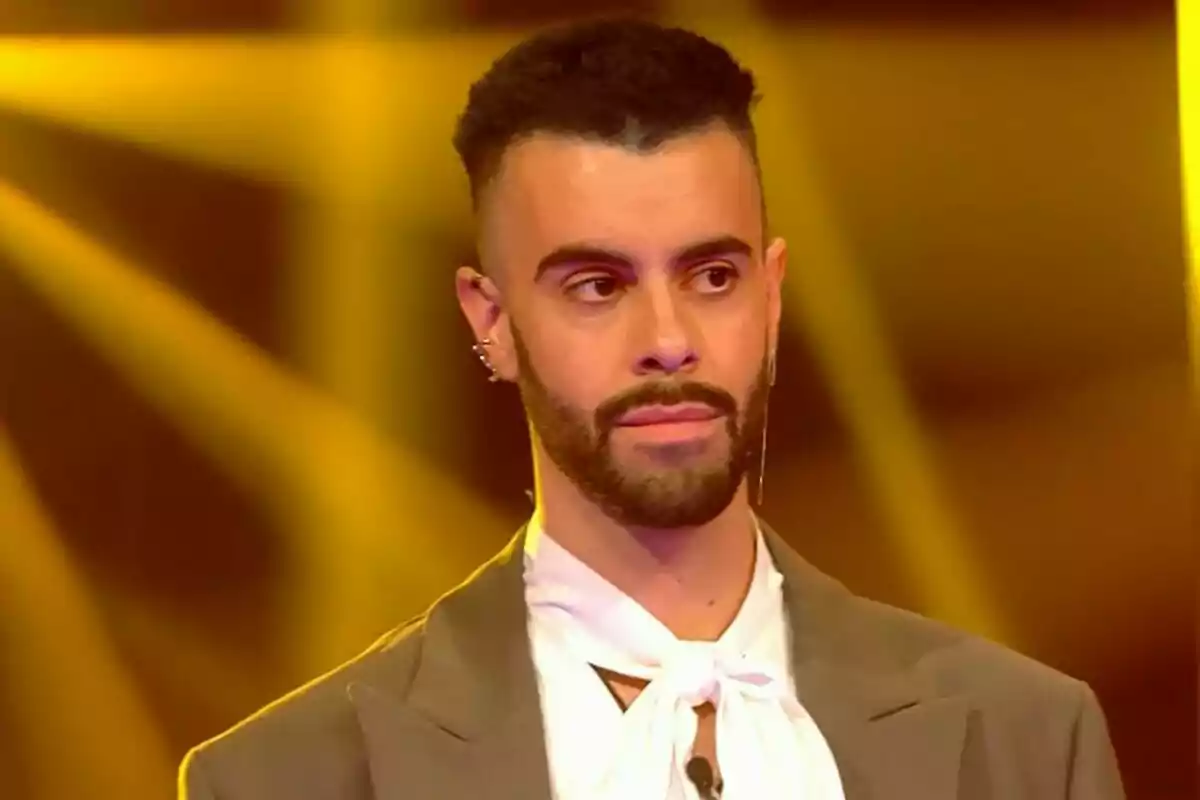 Ruvens con barba y cabello corto lleva un traje gris y una camisa blanca con un lazo en el cuello, iluminado por luces amarillas de fondo en Gran Hermano. Ruvens con barba y cabello corto lleva un traje gris y una camisa blanca con un lazo en el cuello, iluminado por luces amarillas de fondo en Gran Hermano.