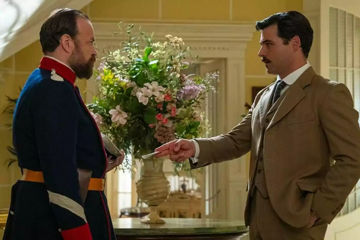 Arturo Sancho i Fran Cants vestits d'època conversen en una habitació elegant amb un gran arranjament floral al fons a La Promesa. Arturo Sancho i Fran Cants vestits d'època conversen en una habitació elegant amb un gran arranjament floral al fons a La Promesa.