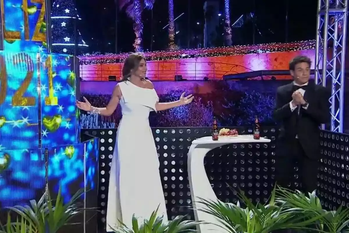 Sandra Barneda y Christian Gálvez en un escenario decorado para la celebración de Año Nuevo 2021, con luces y adornos festivos, una mujer con un vestido blanco y un hombre con un traje negro, junto a una mesa con bebidas y comida. Sandra Barneda y Christian Gálvez en un escenario decorado para la celebración de Año Nuevo 2021, con luces y adornos festivos, una mujer con un vestido blanco y un hombre con un traje negro, junto a una mesa con bebidas y comida.