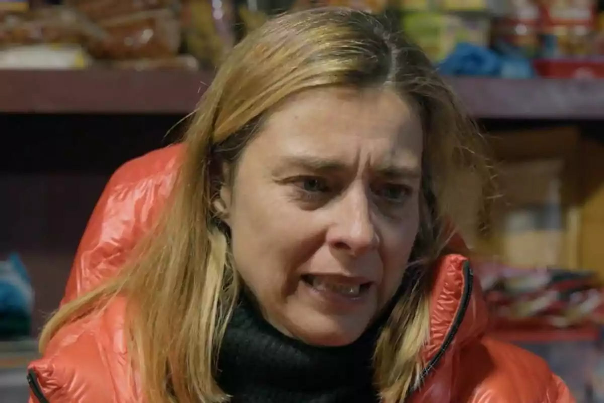 Sandra Barneda con expresión preocupada lleva una chaqueta roja y un suéter negro, con estantes llenos de productos en el fondo en Universo Calleja.
