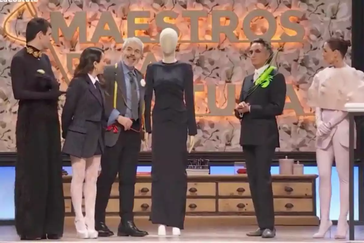 Un grupo de personas elegantemente vestidas está de pie en un escenario con un maniquí en el centro, frente a un fondo que dice Un grupo de personas elegantemente vestidas está de pie en un escenario con un maniquí en el centro, frente a un fondo que dice