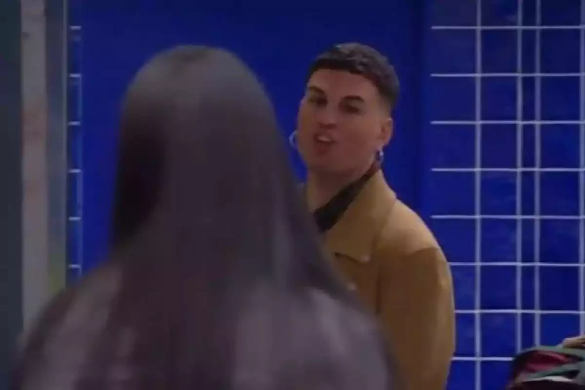 Sergui Aguilera vestido con una chaqueta marrón está haciendo una expresión facial mientras mira a otra persona de espaldas en un entorno con azulejos azules.