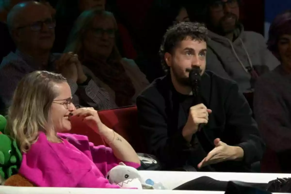 Una dona somrient amb un suèter rosa i Sergio Bezos amb barba sostenint un micròfon, asseguts en un sofà envoltats de públic a La Revuelta. Una dona somrient amb un suèter rosa i Sergio Bezos amb barba sostenint un micròfon, asseguts en un sofà envoltats de públic a La Revuelta.