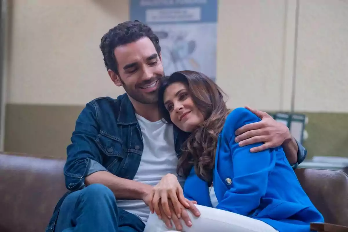 Una pareja sentada en un sofá, el hombre abraza a la mujer mientras ella apoya su cabeza en su hombro y ambos sonríen en la serie Si Nos Dejan Una pareja sentada en un sofá, el hombre abraza a la mujer mientras ella apoya su cabeza en su hombro y ambos sonríen en la serie Si Nos Dejan