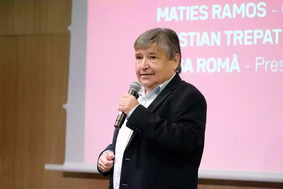Sigfrid Gras de cabells canosos parlant per micròfon davant d’una pantalla amb fons rosa i text blanc de 3Cat.