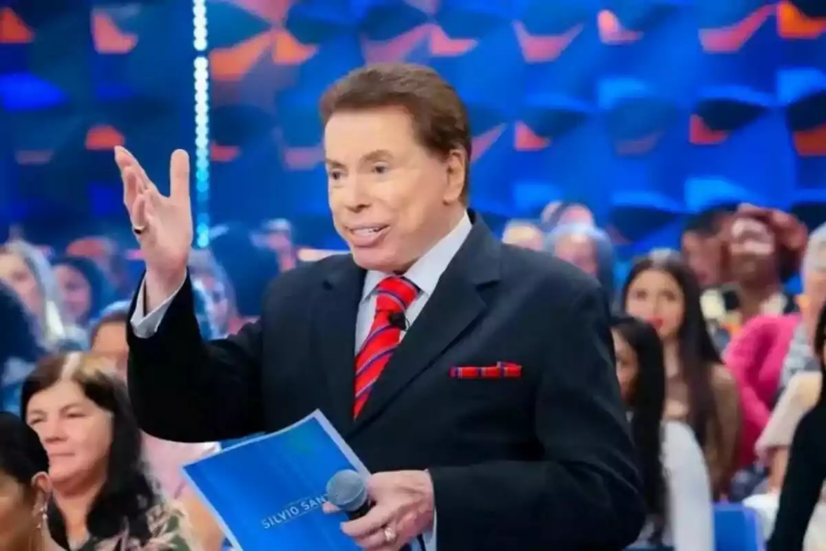Fotografía de Silvio Santos en traje y corbata roja sosteniendo un micrófono y un documento en un escenario con una audiencia al fondo. Fotografía de Silvio Santos en traje y corbata roja sosteniendo un micrófono y un documento en un escenario con una audiencia al fondo.