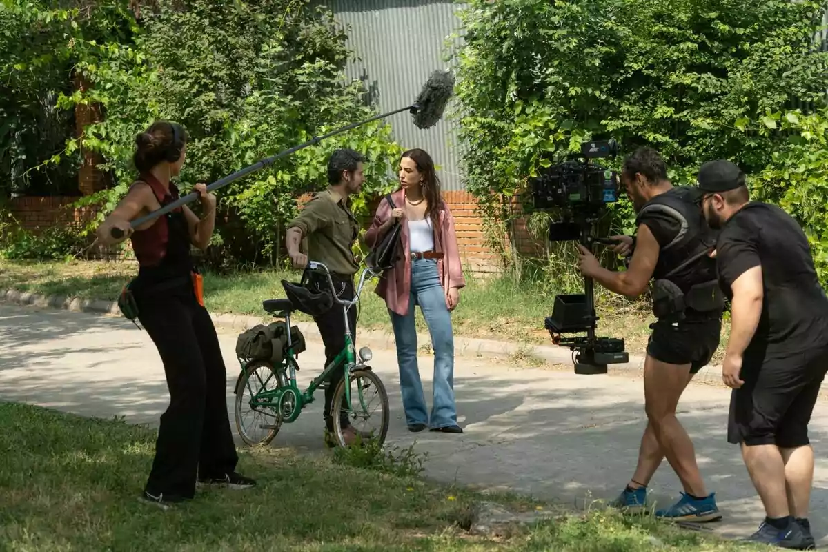 Equip de rodatge gravant una escena a l’aire lliure on una dona i un home amb bicicleta conversen mentre una persona sosté una perxa de micròfon i dos càmeres els enregistren a ‘Sense gluten’.