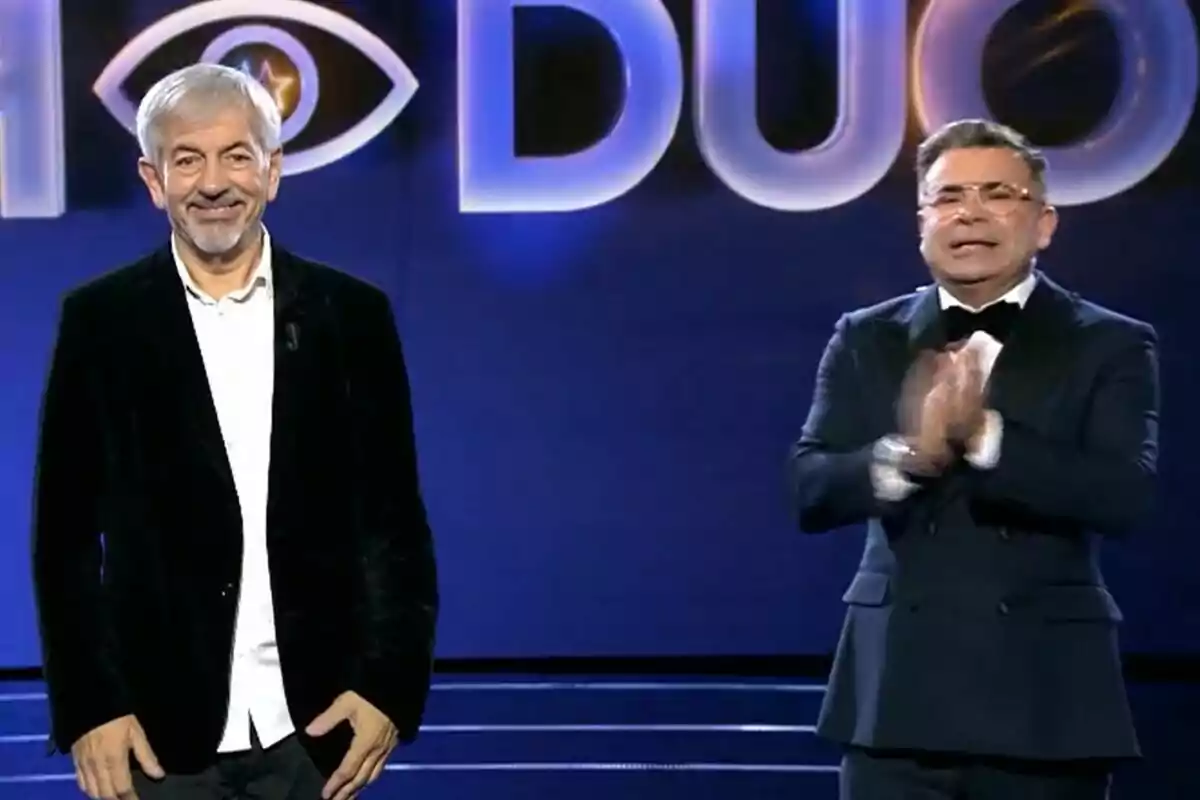 Carlos Sobera y Jorge Javier Vázquez de pie en un escenario con un fondo azul y letras grandes iluminadas de GH DÚO. Carlos Sobera y Jorge Javier Vázquez de pie en un escenario con un fondo azul y letras grandes iluminadas de GH DÚO.