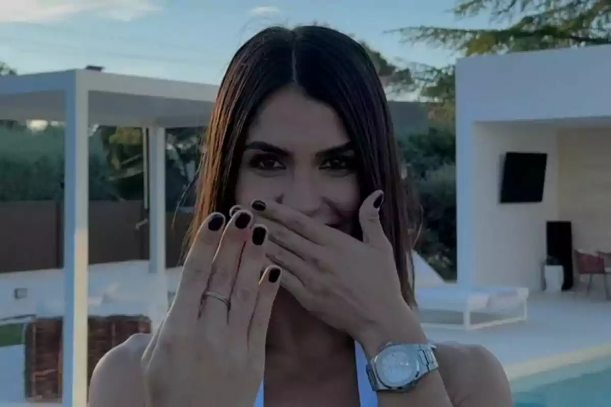 Sofía Suescun sonriente con el cabello oscuro cubre parcialmente su boca con una mano mientras muestra un anillo en la otra mano, en un entorno al aire libre con una piscina y muebles de jardín. Sofía Suescun sonriente con el cabello oscuro cubre parcialmente su boca con una mano mientras muestra un anillo en la otra mano, en un entorno al aire libre con una piscina y muebles de jardín.