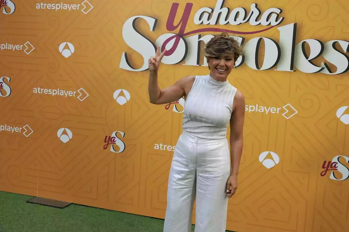 Sonsoles Ónega sonriente vestida de blanco hace el signo de la paz con los dedos frente a un fondo naranja con el logo de Sonsoles Ónega sonriente vestida de blanco hace el signo de la paz con los dedos frente a un fondo naranja con el logo de