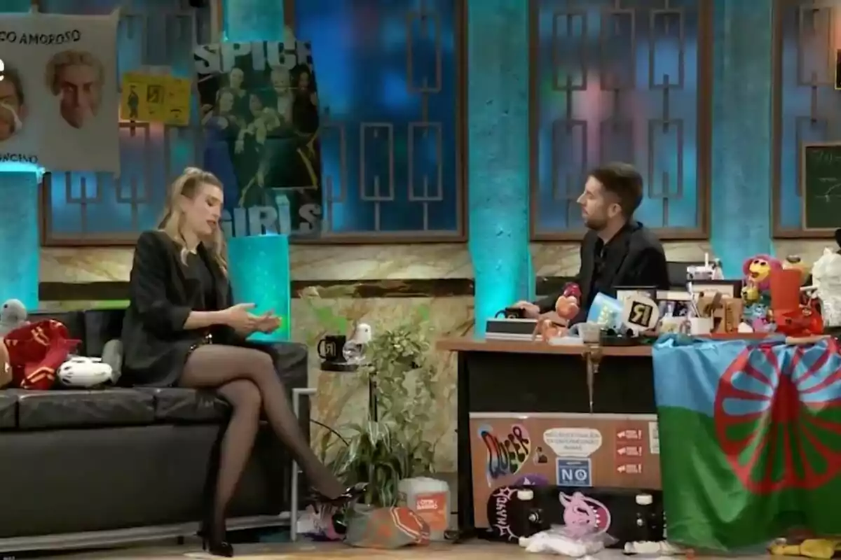 David Broncano y Eva Soriano conversan en un set de televisión decorado con carteles y objetos variados, mientras ella está sentada en un sofá y él detrás de un escritorio lleno de artículos diversos en 'La Revuelta'. David Broncano y Eva Soriano conversan en un set de televisión decorado con carteles y objetos variados, mientras ella está sentada en un sofá y él detrás de un escritorio lleno de artículos diversos en 'La Revuelta'.