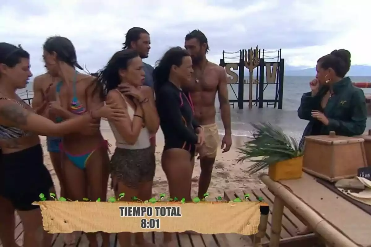 Los concursantes de 'Supervivientes' en la playa, algunas en traje de baño, con un cartel que muestra Los concursantes de 'Supervivientes' en la playa, algunas en traje de baño, con un cartel que muestra