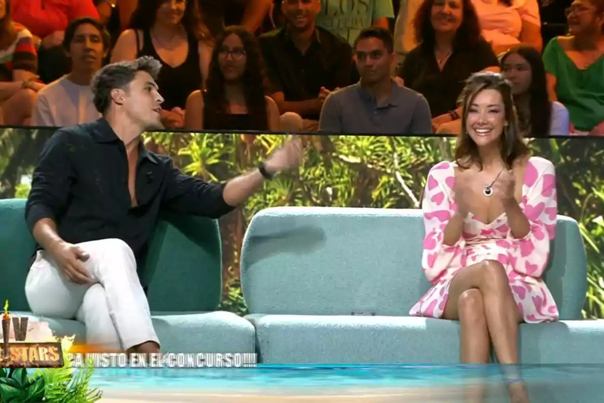 Dos personas están sentadas en un sofá en un programa de televisión, una de ellas aplaude mientras la otra gesticula con la mano, y al fondo se puede ver al público. Dos personas están sentadas en un sofá en un programa de televisión, una de ellas aplaude mientras la otra gesticula con la mano, y al fondo se puede ver al público.