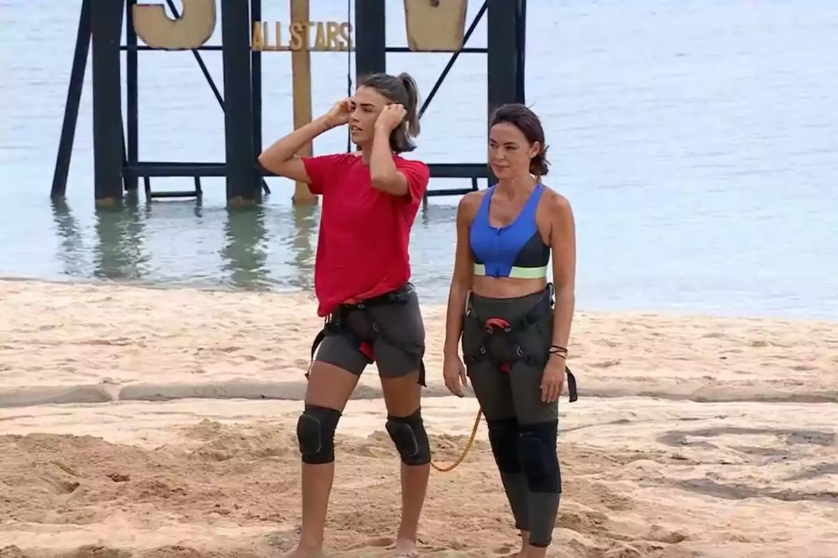 Sofía Suescun y Olga Moreno en la playa con equipo de protección, Sofía con camiseta roja y Olga con top azul, con un cartel de Sofía Suescun y Olga Moreno en la playa con equipo de protección, Sofía con camiseta roja y Olga con top azul, con un cartel de