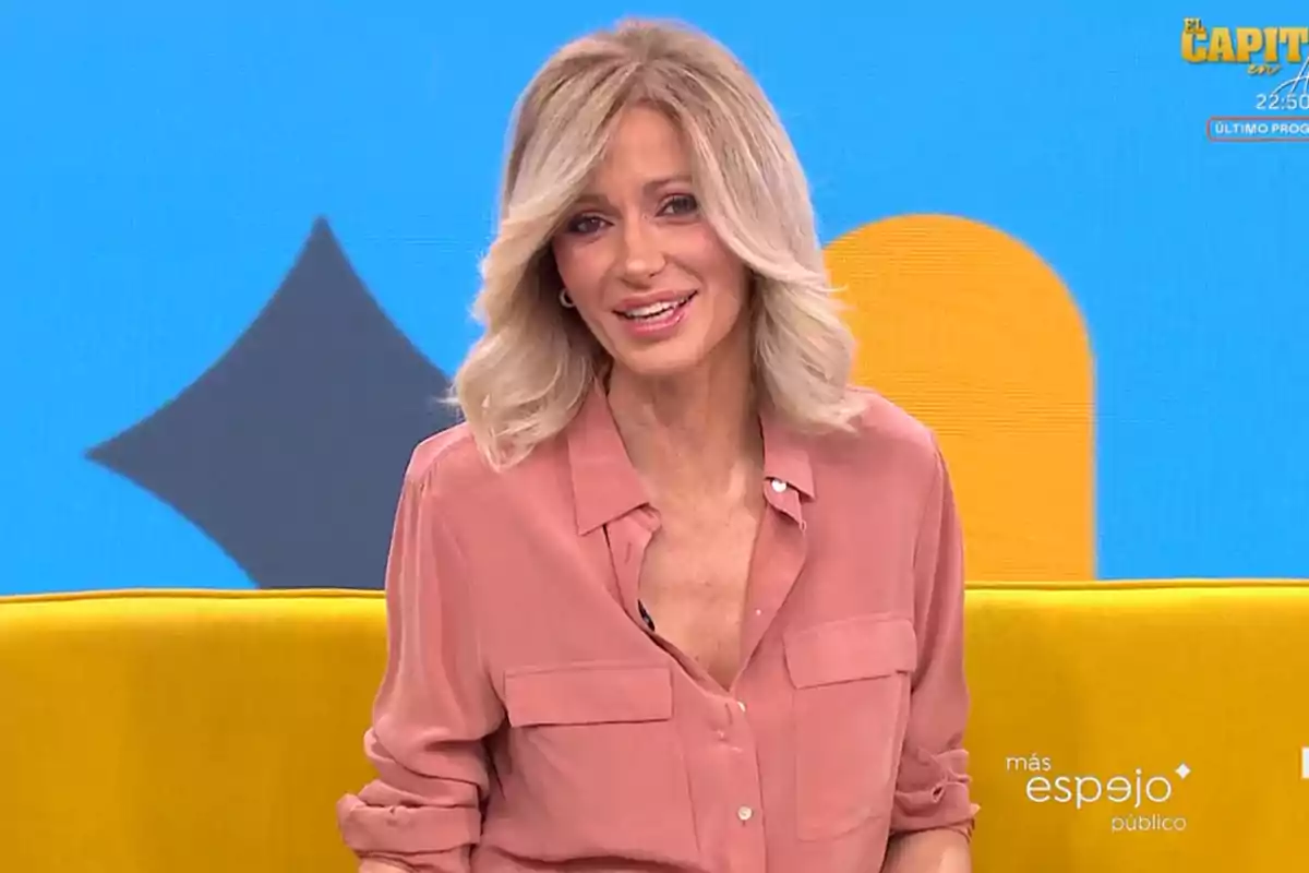 Mujer con cabello rubio y camisa rosa sentada en un set de televisión con fondo azul y amarillo. Mujer con cabello rubio y camisa rosa sentada en un set de televisión con fondo azul y amarillo.