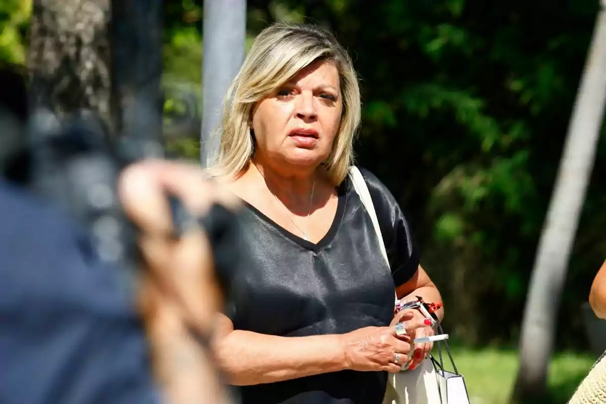 Terelu Campos con una camiseta negra y una bolsa blanca, sosteniendo varios objetos en sus manos, con un fondo de árboles y vegetación. Terelu Campos con una camiseta negra y una bolsa blanca, sosteniendo varios objetos en sus manos, con un fondo de árboles y vegetación.