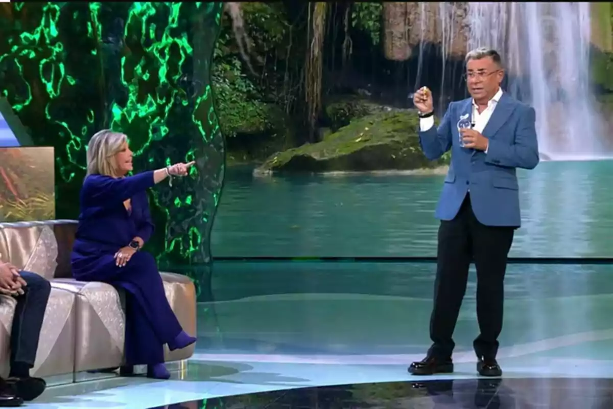 Terelu Campos sentada señala Jorge Javier Vázquez de pie en un set de Supervivientes con un fondo de cascada. Terelu Campos sentada señala Jorge Javier Vázquez de pie en un set de Supervivientes con un fondo de cascada.
