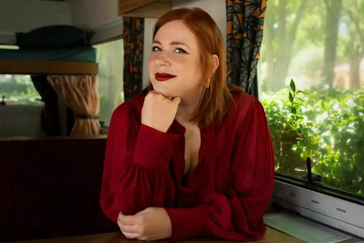 Elísabet Benavent con blusa roja sentada junto a una ventana en el interior de una caravana, apoyando el mentón en su mano y sonriendo suavemente en Toda la verdad de mis mentiras.