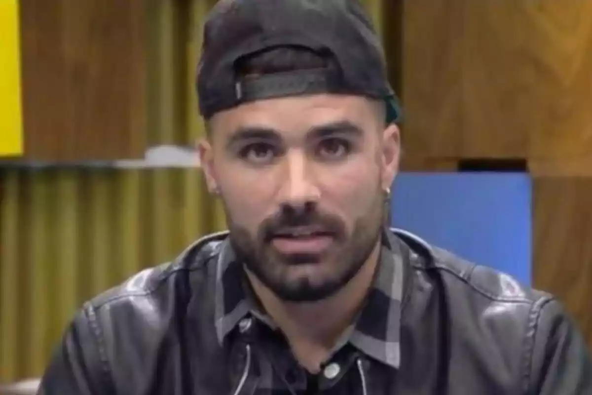 Tutto Durán con barba y gorra al revés, vistiendo una chaqueta de cuero y camisa a cuadros, está mirando hacia la cámara en un entorno interior con fondo de madera y cortinas.
