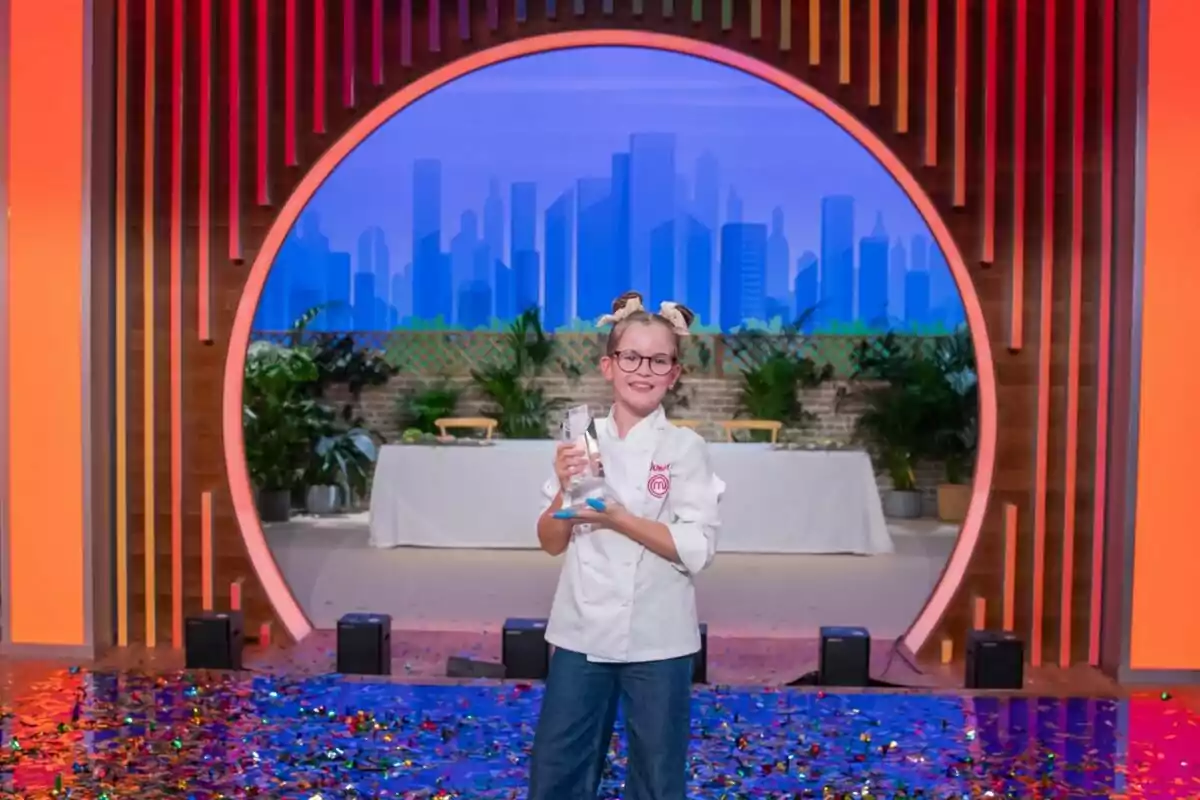 Valentina amb uniforme de xef sosté un trofeu de MasterChef Junior en un escenari decorat amb confeti i un fons de ciutat il·luminada. Valentina amb uniforme de xef sosté un trofeu de MasterChef Junior en un escenari decorat amb confeti i un fons de ciutat il·luminada.