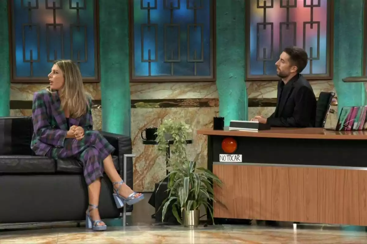 Valeria Ros con traje de cuadros morados y verdes está sentada en un sofá negro, mientras David Broncano con camisa negra está sentado detrás de un escritorio de madera con un letrero que dice Valeria Ros con traje de cuadros morados y verdes está sentada en un sofá negro, mientras David Broncano con camisa negra está sentado detrás de un escritorio de madera con un letrero que dice