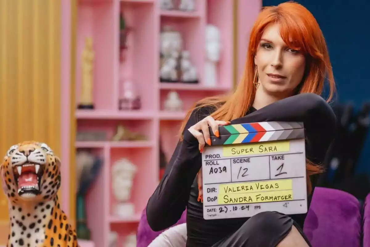 Valeria Vegas con cabello rojo sostiene una claqueta de cine en un set colorido con una figura de leopardo al lado. Valeria Vegas con cabello rojo sostiene una claqueta de cine en un set colorido con una figura de leopardo al lado.