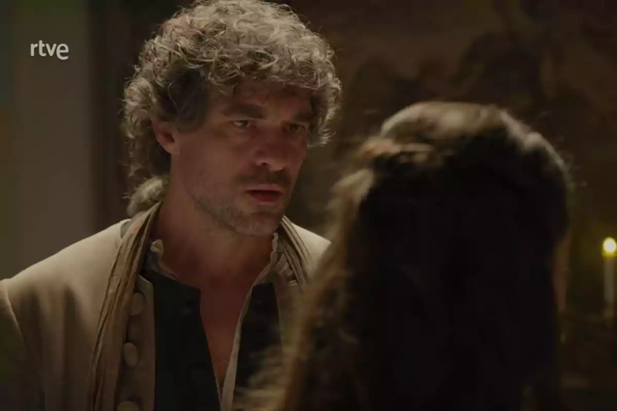 Bernardo amb cabell arrissat i expressió seriosa mira una persona d'esquena en una escena il·luminada per una espelma. Captura de 'Valle Salvaje'.