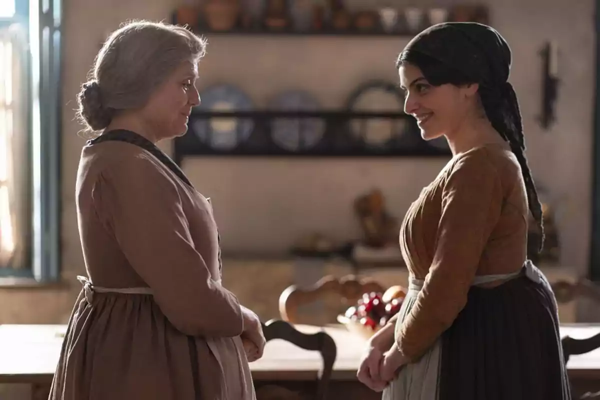 Raimunda y Luisa vestidas con ropa de época se miran y sonríen en una cocina iluminada por la luz del día. Captura de 'Valle Salvaje' Raimunda y Luisa vestidas con ropa de época se miran y sonríen en una cocina iluminada por la luz del día. Captura de 'Valle Salvaje'