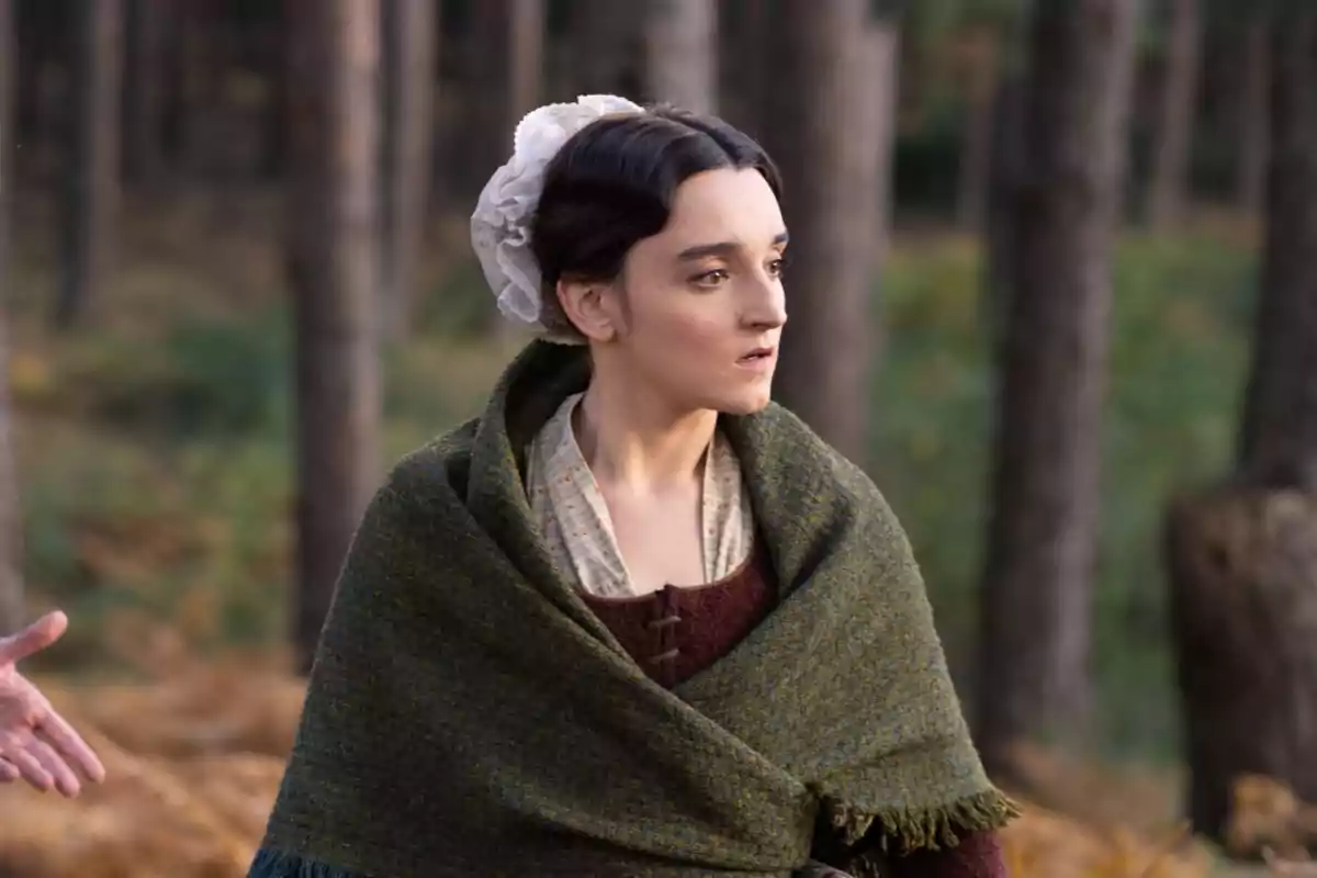 Matilde vestida con ropa de época y un chal verde oscuro está en un bosque, mirando hacia un lado con expresión seria. Captura de 'Valle Salvaje' Matilde vestida con ropa de época y un chal verde oscuro está en un bosque, mirando hacia un lado con expresión seria. Captura de 'Valle Salvaje'