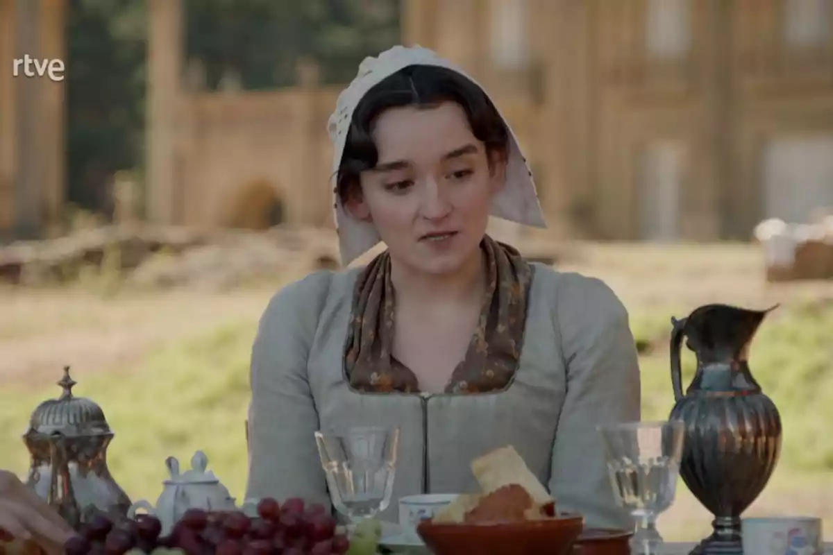 Matilde vestida amb roba d'època i un mocador al cap està asseguda en una taula exterior amb menjar i beguda, en un entorn campestre amb un edifici antic de fons. Captura de 'Valle Salvaje'